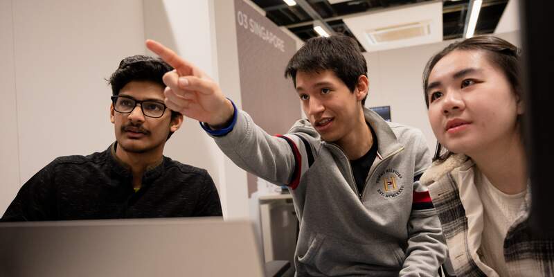 Two students looking at a screen. A third student is sat between and pointing at the screen